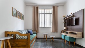 A compact living space with a wooden sofa, a small table, two round stools, a TV mounted on the wall, and a window with curtains.