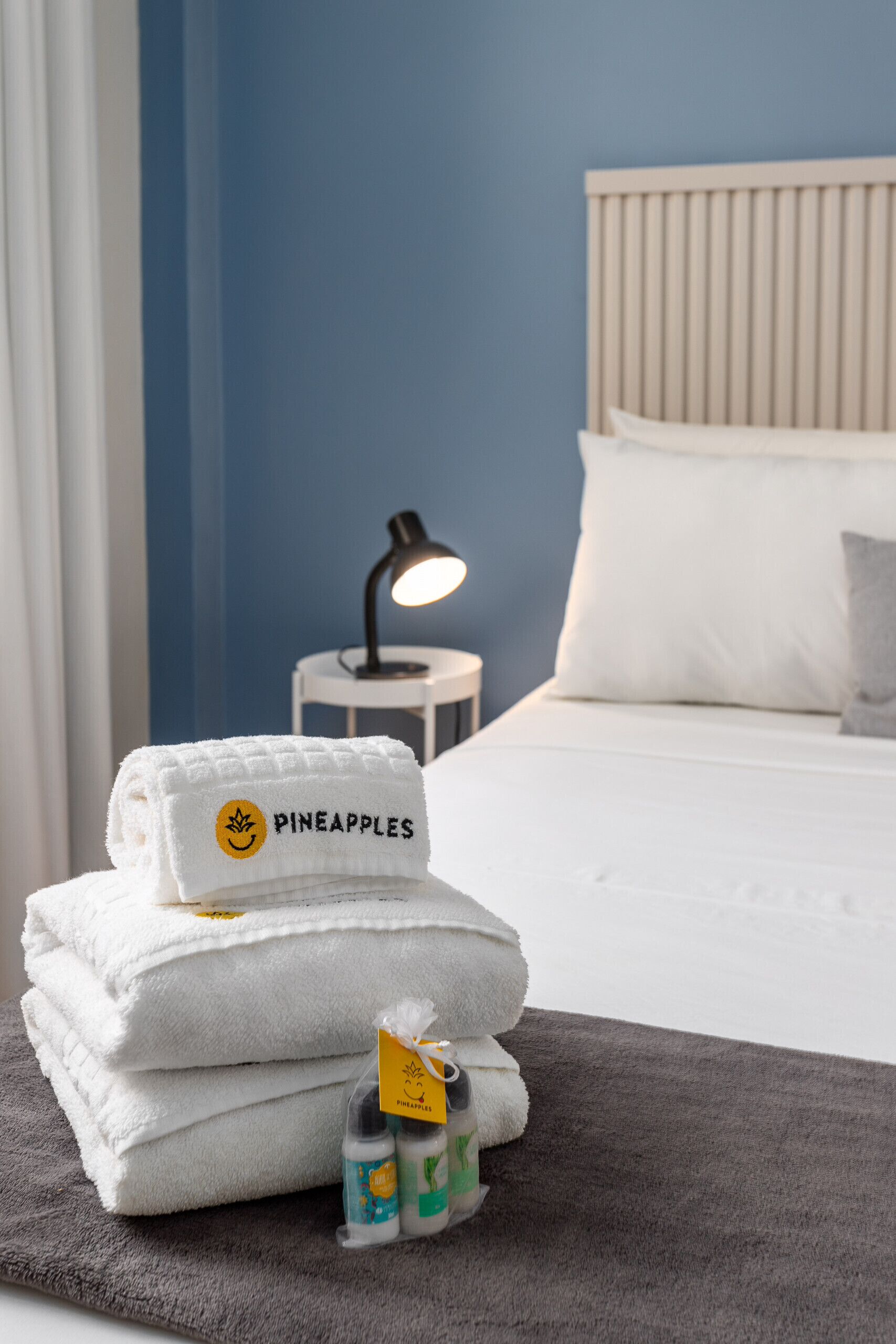 A neatly made bed with white linens and a pillow, a bedside lamp, and a stack of folded towels with a Pineapples logo.