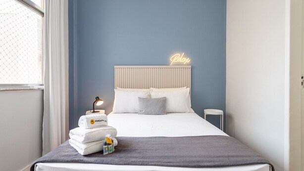 A neatly made bed with a grey blanket and pillows, a small table with a lamp and toiletries, and a neon sign on the wall. - Near the underground and 20 minutes from Maracanã | CV410 (Rio de Janeiro)