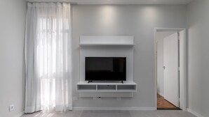 A modern living room with a flat-screen TV mounted on a white wall, a large window with sheer curtains, and a door leading to another room. - Near the underground and 20 minutes from Maracanã | CV410 (Rio de Janeiro)
