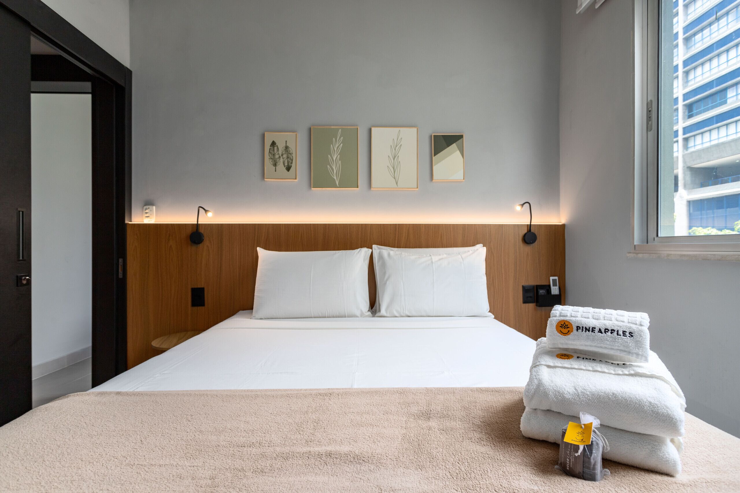Una habitación de hotel con una cama, almohadillas blancas, un cabecero de madera y cuatro láminas enmarcadas con motivos botánicos en la pared.