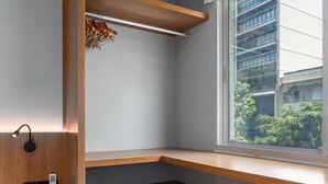 A corner room with built-in wooden shelving, a small stool, a wall-mounted light, and a window with a view of buildings and greenery. - 5 minutes from Leblon Beach | AP302 (Rio de Janeiro)
