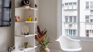 A modern living room with a white sofa, a glass coffee table, a white rocking chair, a white shelving unit with decorative items, and a large window with a view of buildings. - For families, 3 minutes from Copacabana beach | SL501 (Rio de Janeiro)