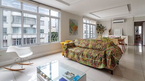 A modern living room with a colorful sofa, a white rocking chair, a glass coffee table, and large windows offering a view of the city. - For families, 3 minutes from Copacabana beach | SL501 (Rio de Janeiro)