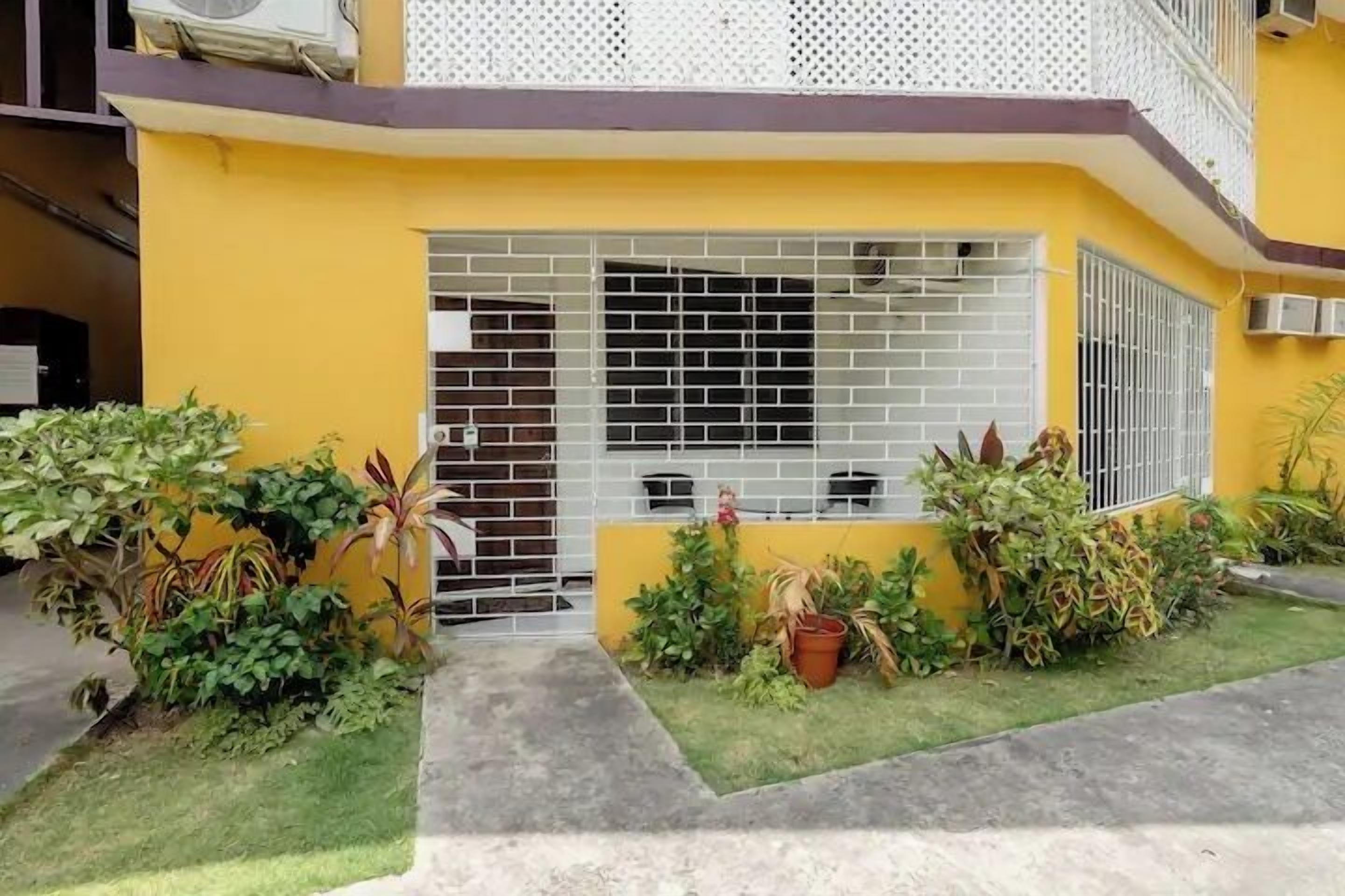 Un bâtiment jaune avec une entrée fermée par un portail, un petit jardin agrémenté de plantes en pot et une fenêtre avec des barreaux en métal.