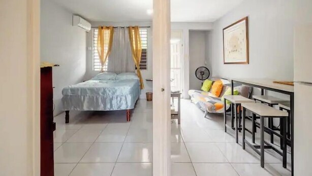 A compact room with a bed, a small seating area with cushions, a table, and a fan. - Camperdown Place (Kingston)