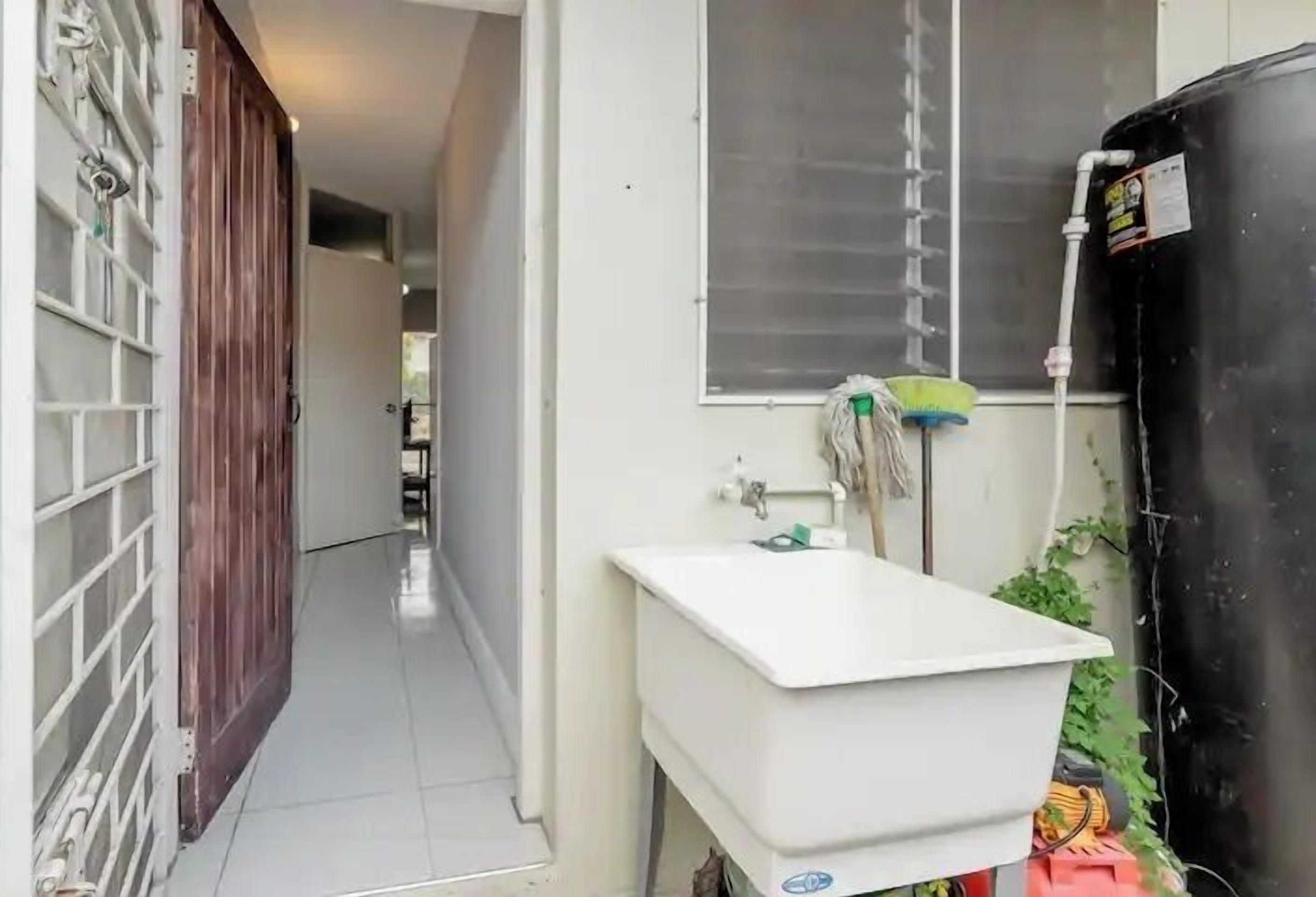 A narrow hallway with a white sink, a window with blinds, and a black water tank.