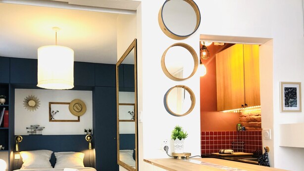 Une chambre moderne avec un lit, des lampes de chevet et un grand miroir. On aperçoit au fond une petite kitchenette équipée d’une cuisinière et d’un luminaire suspendu.