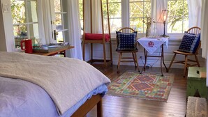 Ein gemütliches Schlafzimmer mit Bett, Schaukelstuhl, kleinem Tisch mit Lampe und großen Fenstern mit Vorhängen.