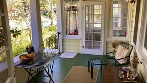 Eine gemütliche Veranda mit Tisch und Stühlen, ein Fenster mit Blick auf Grünflächen und eine Tür, durch die ein Haustier zu sehen ist.