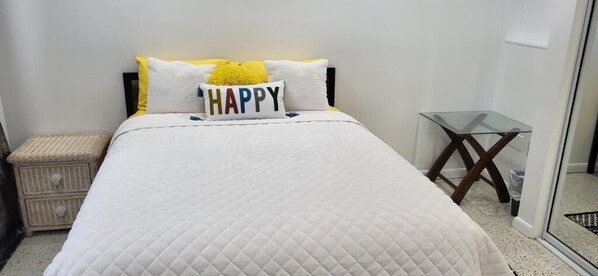 A neatly made bed with a white quilted bedspread and pillows, including one with the word "HAPPY" on it.