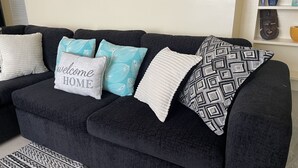 Un sofá de color oscuro con varios cojines, incluyendo un cojín que dice 'bienvenido a casa', un cojín con estampado geométrico y un cojín blanco con borde fruncido.
