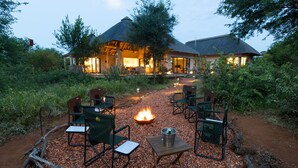 An outdoor seating area with a fire pit, surrounded by chairs and tables, leading to a building with a thatched roof. - Villa Bushman: Wonderful luxury and attractive villa (Hoedspruit)