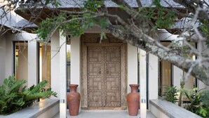 A traditional wooden door with intricate carvings, flanked by two large terracotta pots, leading to a building with a thatched roof and a stone pathway.
