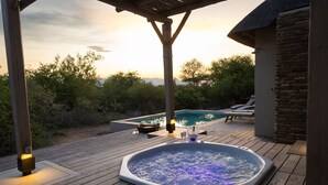 A hot tub on a wooden deck with a pool in the background, surrounded by greenery.