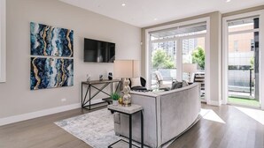 Een moderne woonkamer met een grijze bank, een salontafel en een aan de muur bevestigde televisie. Aan de wand hangen abstracte schilderijen en een raam biedt uitzicht op het omliggende landschap.