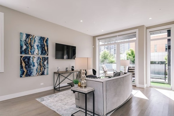 A modern living room with a grey sofa, a coffee table, and a TV mounted on the wall. There are abstract paintings on the wall and a window providing a view of the outside. - 2 Newly Built Luxury Condos with Private Roofdeck! (Chicago)