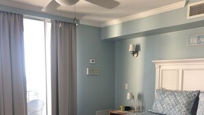 A bedroom with a bed, a ceiling fan, a window with curtains, a bedside table with a lamp, and a wall sign.
