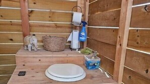 A wooden outdoor toilet with a white seat, a small trash bin, a tissue roll, a cleaning spray bottle, and a decorative elephant figurine. - Glamping Tent w Queen Size Bed & Incredible Views (Terlingua)