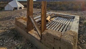 A rustic outdoor setting with a wooden structure, possibly a fire pit, and a white tent in the background. - Glamping Tent w Queen Size Bed & Incredible Views (Terlingua)