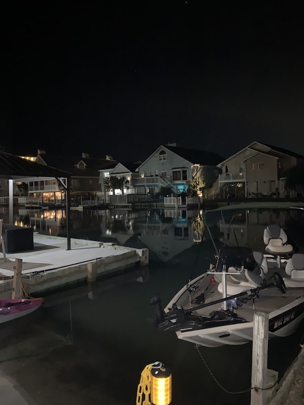 Una marina de noche con barcos amarrados, agua tranquila y casas al fondo.