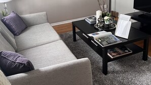 A living room with a grey sofa, a black coffee table with magazines and a framed picture, a television, and wall art.