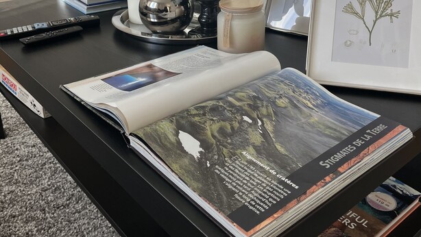 A desk with a magazine, a framed botanical illustration, a candle, and a decorative vase.
