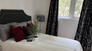 A neatly made bed with white bedding, a red pillow, and a plant on the nightstand.