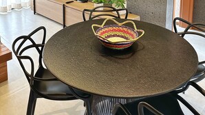 A modern dining area with a round table, black chairs, a woven pendant light, and a television on the wall. - Apartamento 02 Quartos - São Miguel dos Milagres (São Miguel dos Milagres)