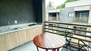 A modern outdoor kitchen with a round wooden table and chairs, a built-in sink, and a view of a residential building. - Apartamento 02 Quartos - São Miguel dos Milagres (São Miguel dos Milagres)