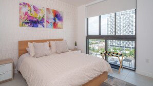 A modern bedroom with a large bed, two abstract paintings, a wooden headboard, and a view of a cityscape through a large window. - Stylish 2BR Condo. Walk to Restaurants & Shops (Doral)