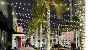 A nighttime street scene with palm trees and string lights. - Stylish 2BR Condo. Walk to Restaurants & Shops (Doral)