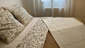 A neatly made bed with patterned pillows and a white throw blanket, a window with sheer curtains, and a brick wall in the background. - Appartement (Damery)