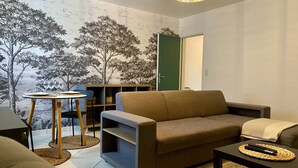 A living room with a sofa, a coffee table, and a small table with wine glasses. There is a bookshelf and a door in the background. - Appartement (Damery)