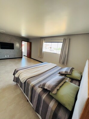 A spacious bedroom with a large bed, a flat-screen TV mounted on the wall, and a window with curtains.