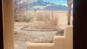 A view through a window of a room with a stone wall, a wooden bench, and a view of a mountainous landscape. - Taos Mountain Views Cozy Home Special Rates (El Prado)