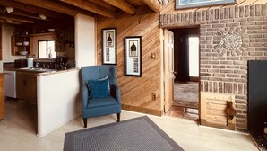 A rustic interior with exposed wooden beams, a fireplace, a kitchen area, and a blue armchair. - Taos Mountain Views Cozy Home Special Rates (El Prado)