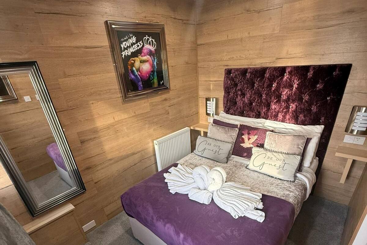 A modern bedroom with a wooden wall panel, a large bed with a purple bedspread, a headboard with a floral design, and a poster on the wall.