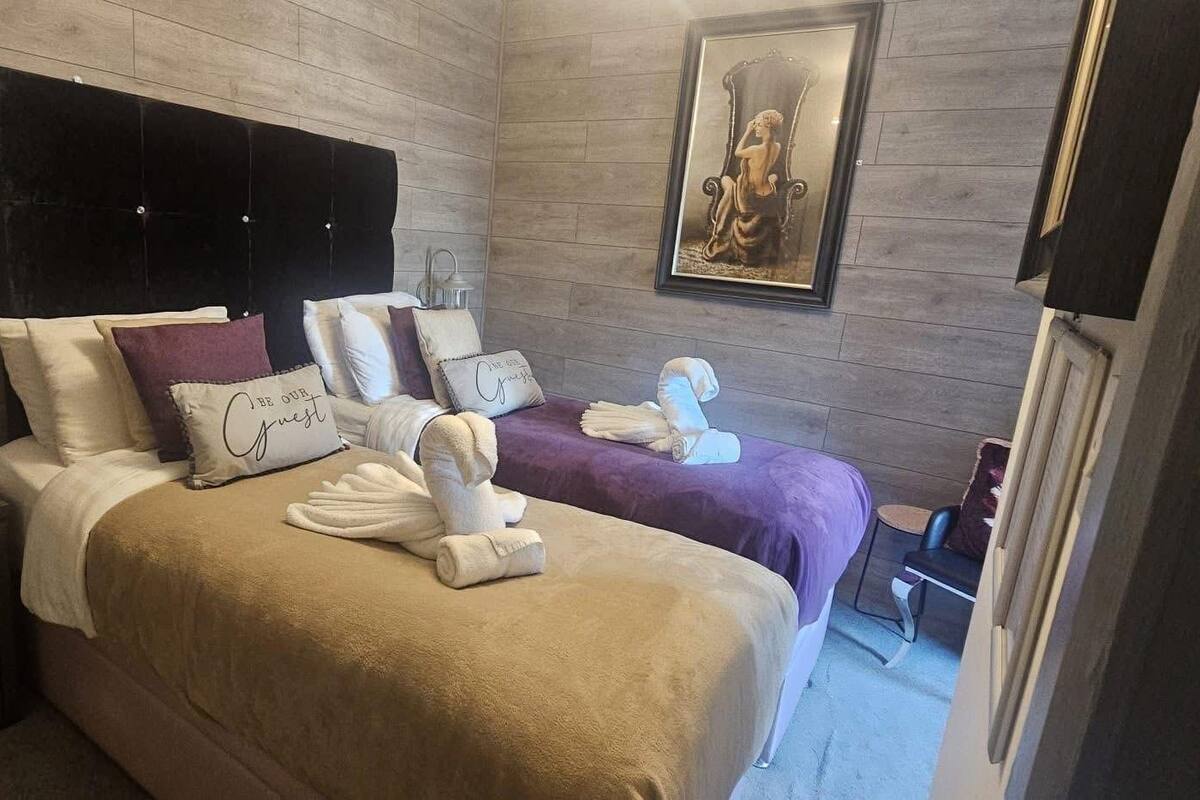 A hotel room with two beds, a headboard, a nightstand, a wall-mounted lamp, a framed picture, and a chair.
