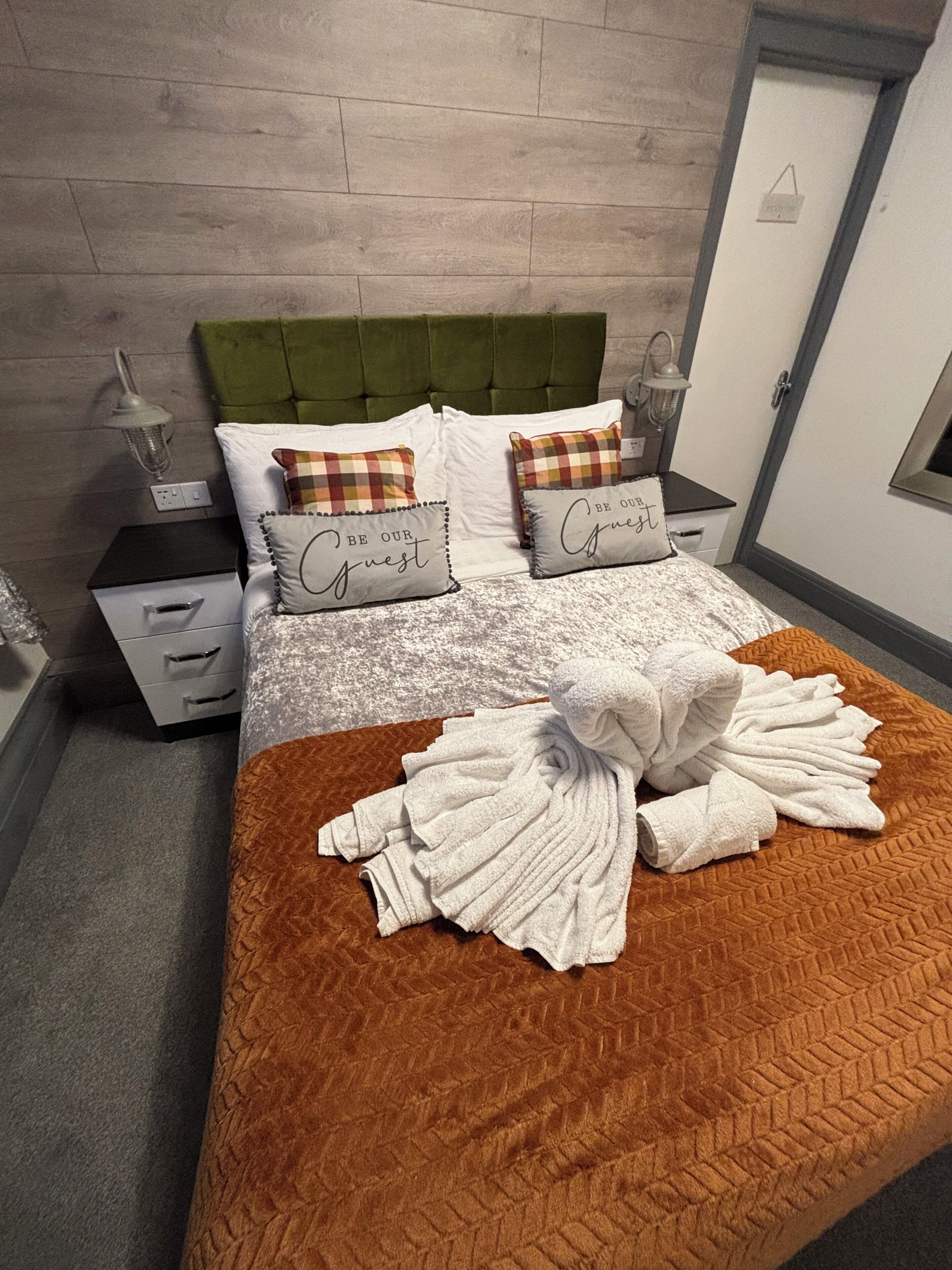 A neatly made bed with a brown bedspread and white towels, a green upholstered headboard, and two bedside tables with lamps.