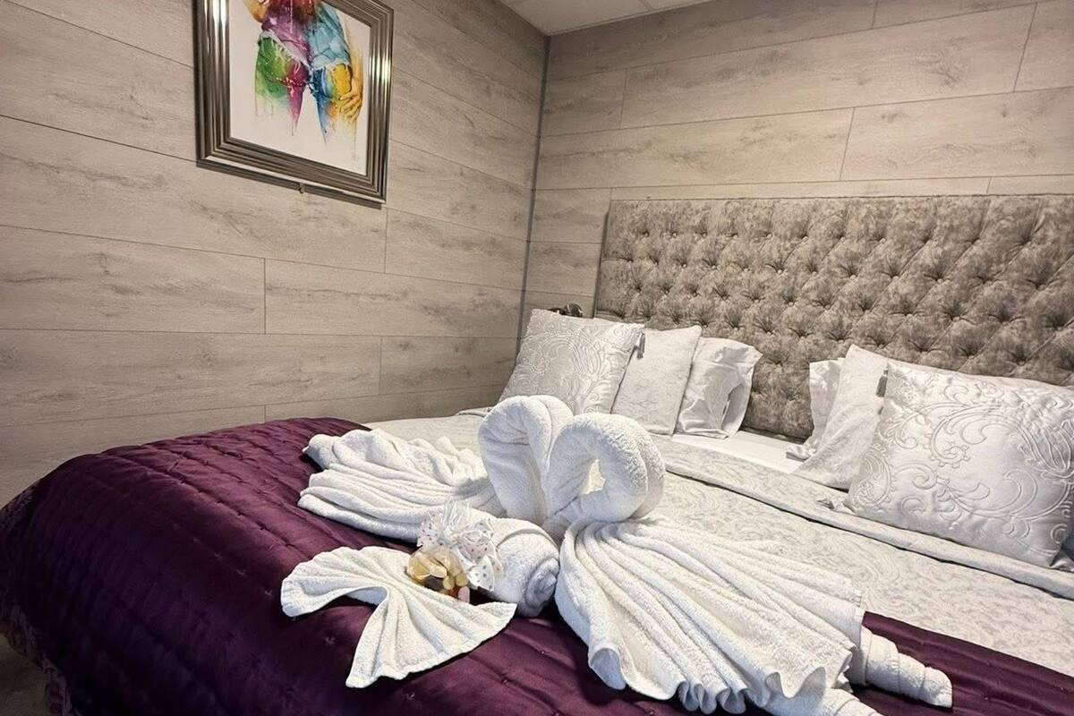 A hotel room with a bed, a headboard, a framed picture on the wall, and a purple bedspread.