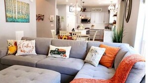A modern living room with a grey sectional sofa, a matching ottoman, and decorative pillows. A kitchen is visible in the background with white cabinets and a 'GATHER' sign. - Beautiful Modern Farmhouse
 (Keizer)