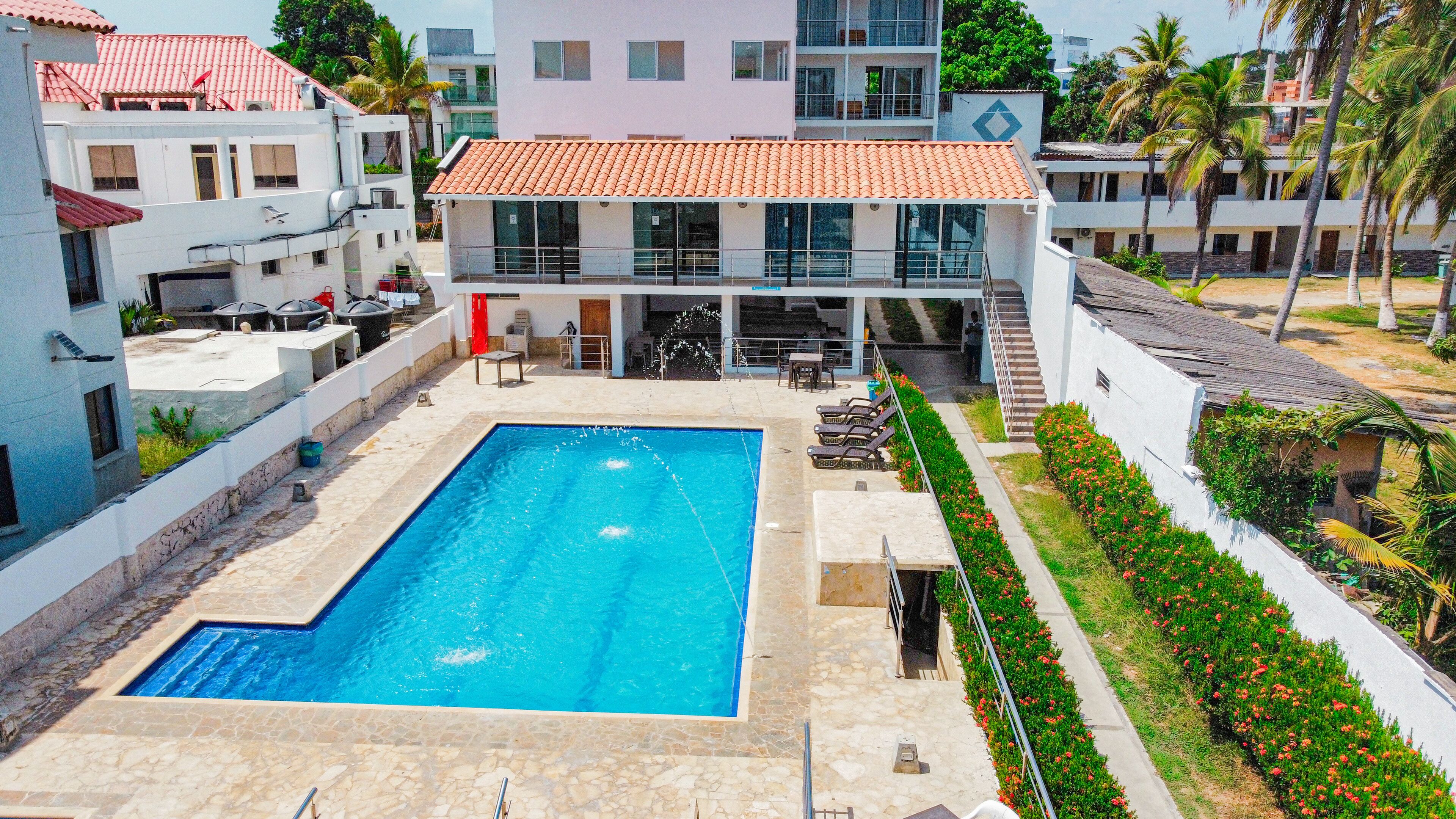 A swimming pool with a tiled deck, surrounded by a garden with red flowers and palm trees.