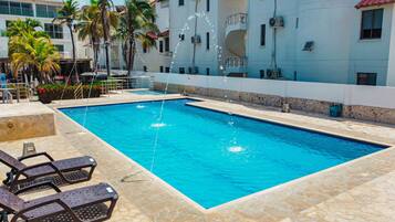 Una piscina rectangular con agua azul cristalina, rodeada de baldosas de piedra. Hay sillas de descanso y una pequeña zona junto a la piscina con una palmera y flores.