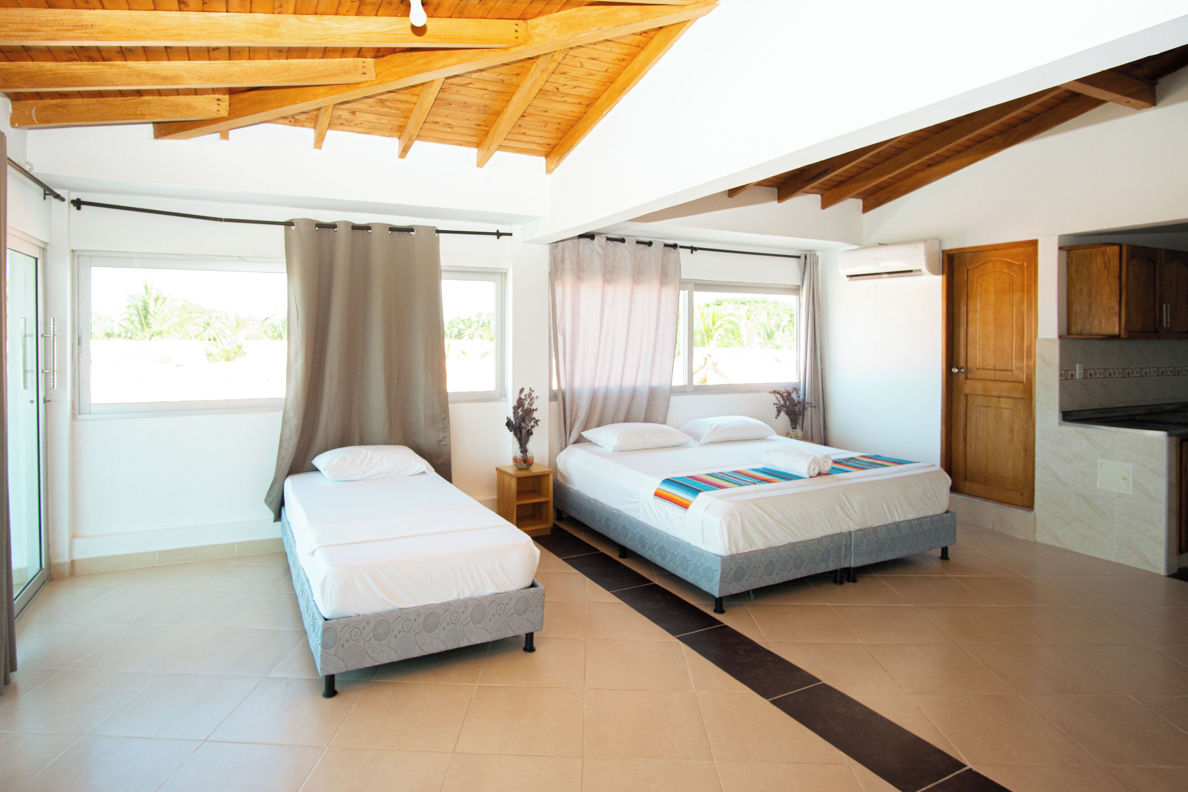 A modern hotel room with two beds, a wooden ceiling, large windows, and a kitchenette.
