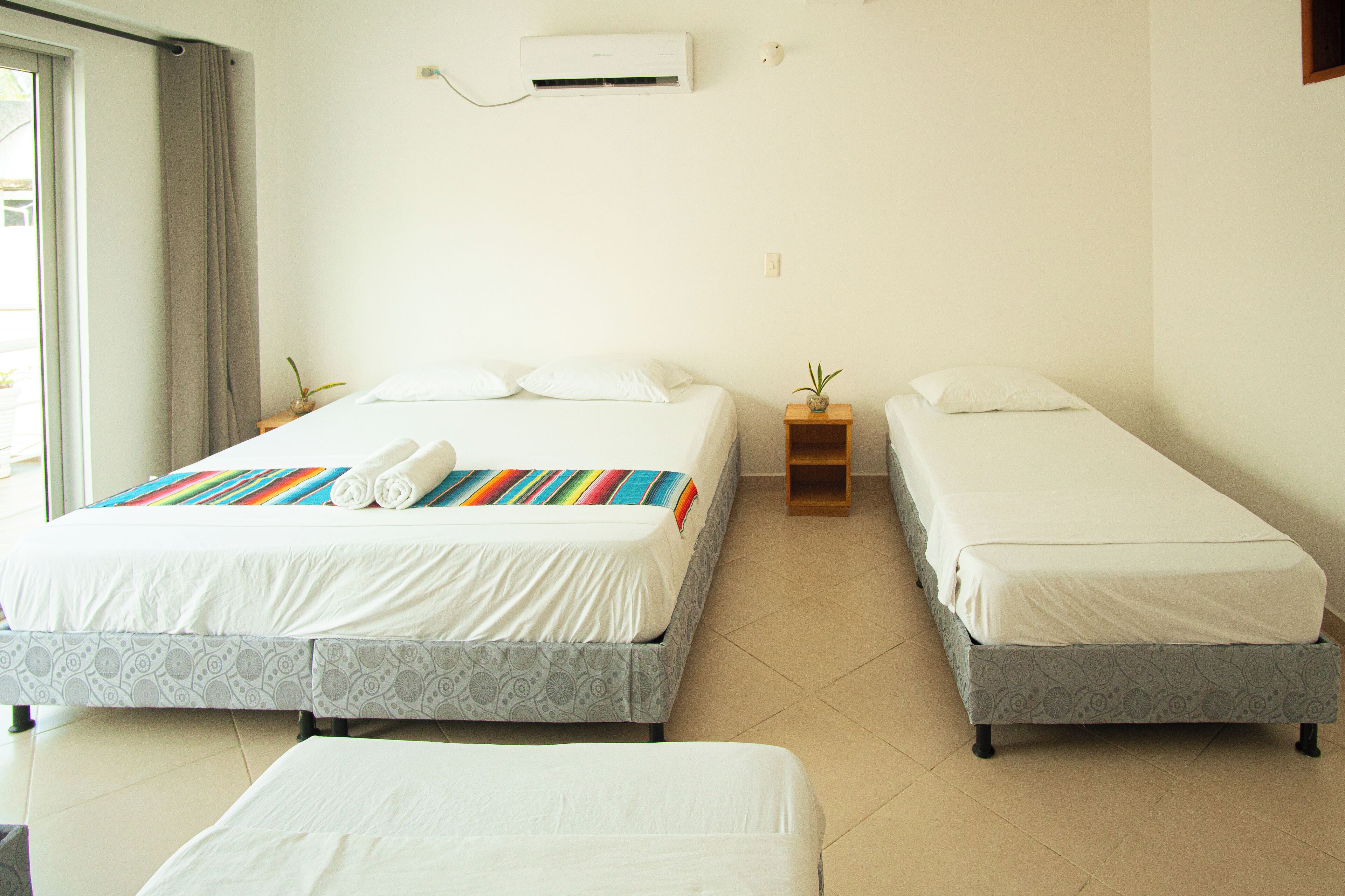 A hotel room with two single beds, a small bedside table with a plant, and a large window with curtains.