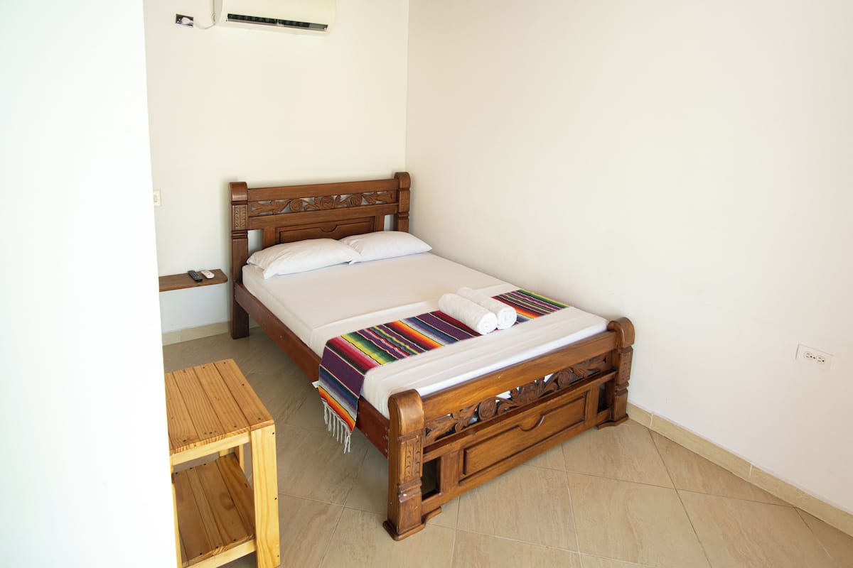Un lit bien fait, avec une tête de lit en bois, une petite table en bois à côté, et un mur sobre agrémenté d’une prise de courant.