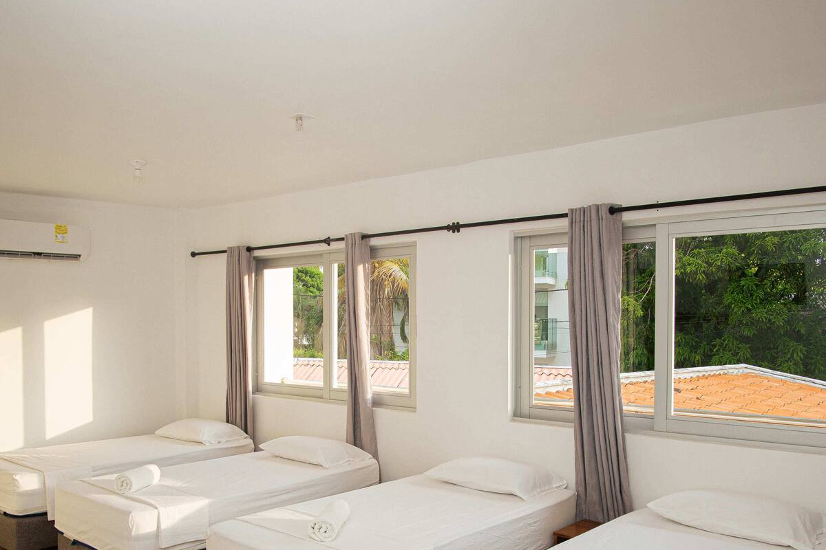 Une chambre d’hôtel avec quatre lits simples, une grande fenêtre avec des rideaux et un sol carrelé.
