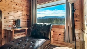 A wooden cabin room with a black leather chair, a small table with a lamp, and a window offering a mountain view. - 8 King Beds, 3 Levels, Hot tub, Pool table, Bar, Wood Soaker, Vaulted with VIEWS (Gatlinburg)