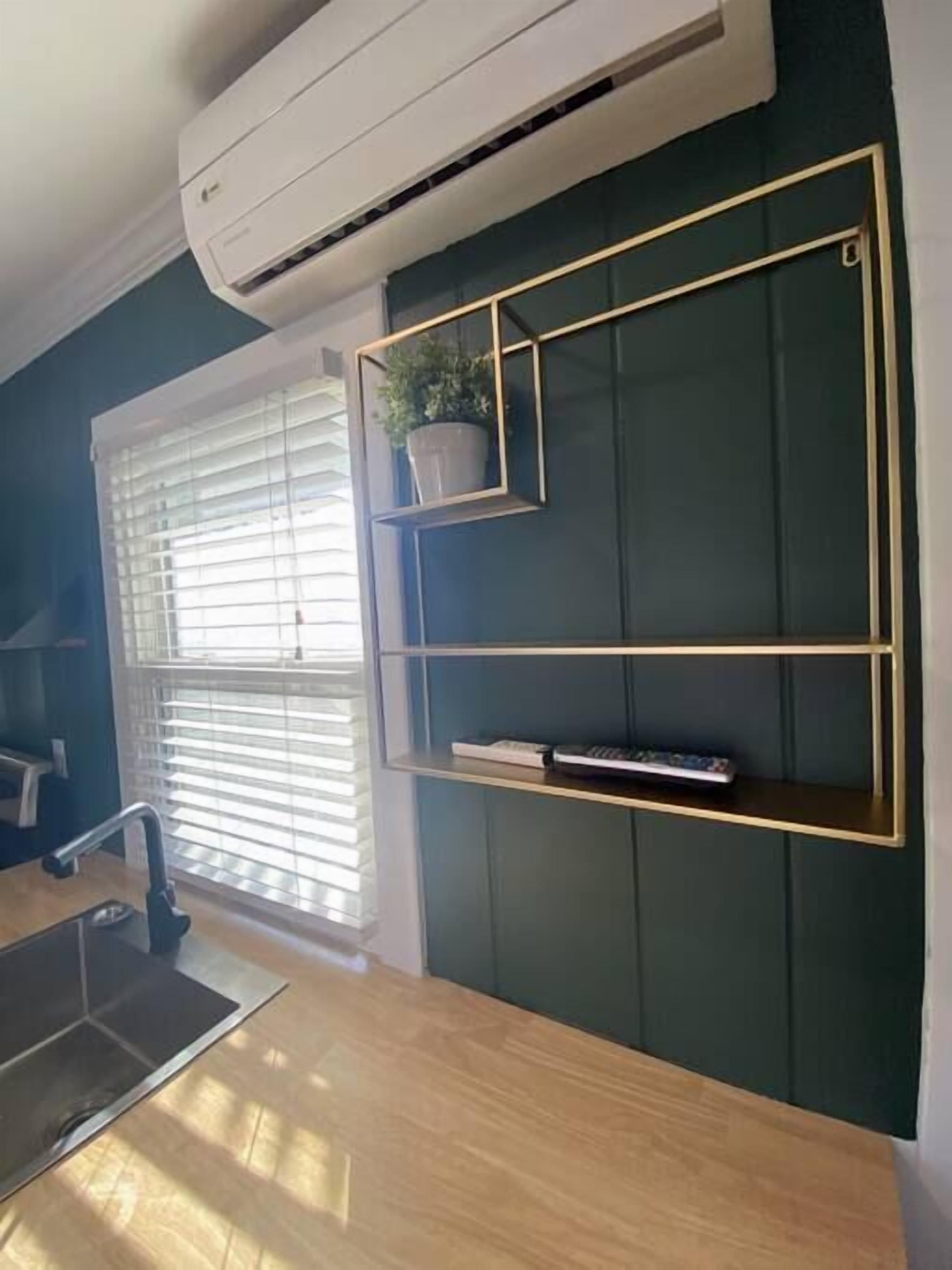A modern kitchen with a wooden countertop, an air conditioning unit, a window with blinds, and a wall-mounted shelf with a potted plant.